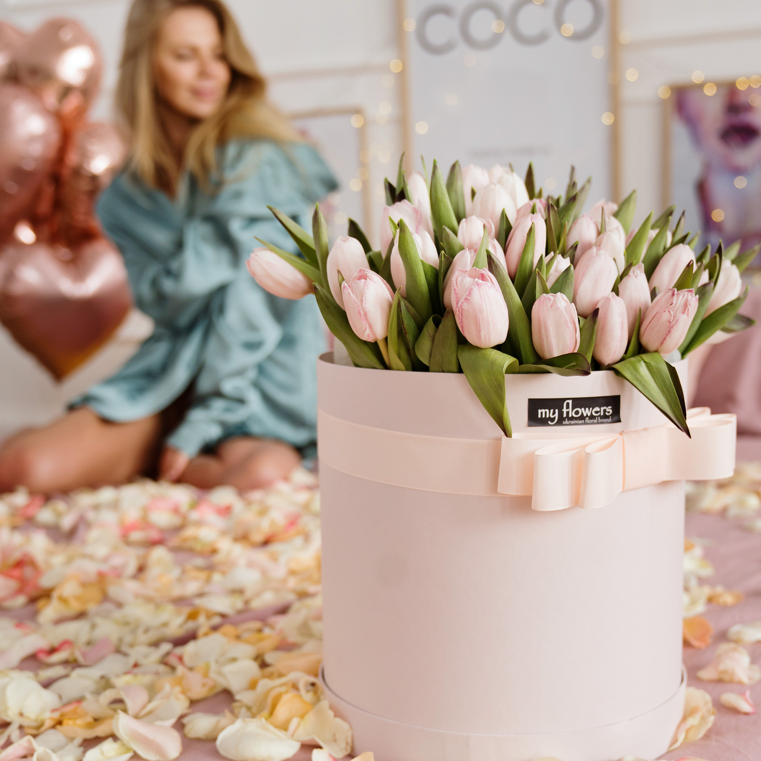 A round pale pink flower box filled with light pink tulips, tied with a matching ribbon and bow. A woman in a blue robe sits blurred in the background near rose gold balloons, with scattered flower petals on the floor.