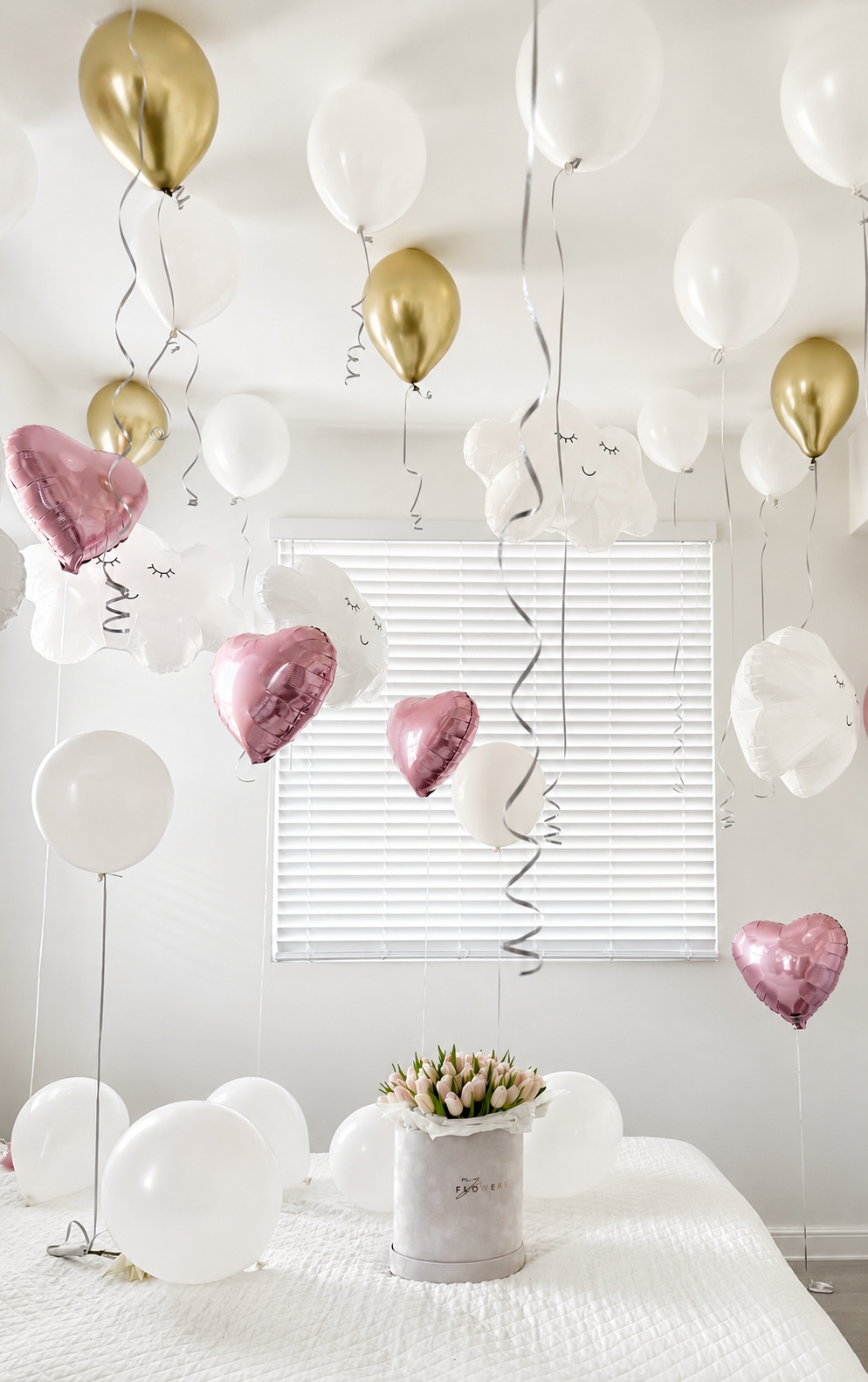 A festive room decorated with numerous white, metallic gold, and metallic pink heart-shaped and round balloons floating around a bed with a bouquet.