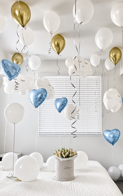 A decorated bedroom with white and gold balloons, including metallic heart-shaped balloons and cloud-shaped balloons, hanging from the ceiling and arranged around a bed with a small floral box in the center.