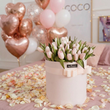 A round pale pink flower box filled with light pink tulips, tied with a matching ribbon and bow. Rose gold balloons, with scattered flower petals on the floor.