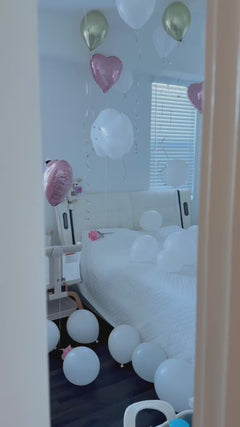 A festive room decorated with numerous white, metallic gold, and metallic pink heart-shaped and round balloons floating around a bed with a bouquet.